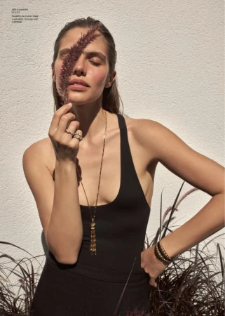 Fashion model in a black tank top holding ornamental grass over one eye against a sunlit white textured wall.