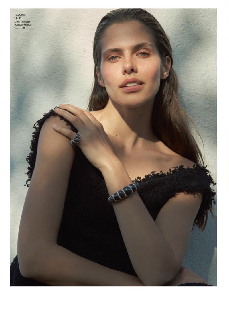 A woman wearing a black textured dress and Cartier bracelet resting her hand on her shoulder against a sunlit wall.