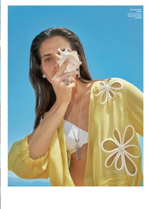 Model holding a white seashell to her face wearing a sheer yellow floral applique blouse against a clear blue sky.