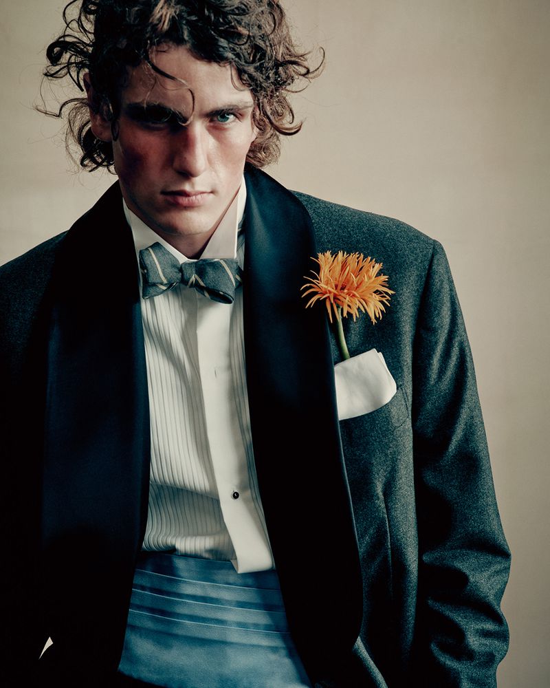 Artjom Zagrebaylov posing in a dark tuxedo jacket with a striped bowtie, blue cummerbund, and an orange flower boutonniere.