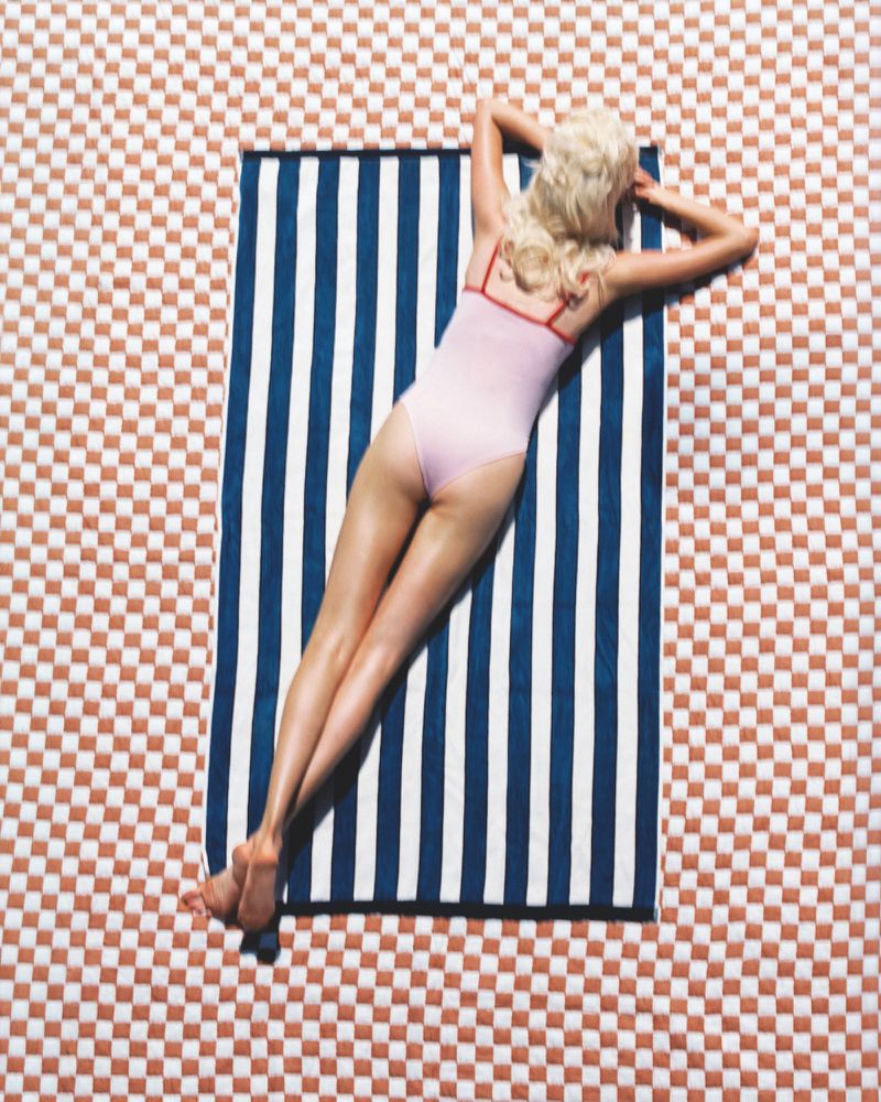 Overhead view of a blonde model lying face down on a blue and white striped towel over a checkered pattern surface.