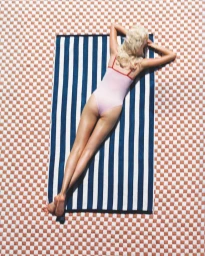 Overhead view of a blonde model lying face down on a blue and white striped towel over a checkered pattern surface.