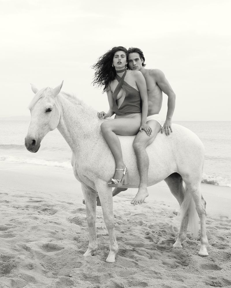 Black and white portrait of an adult couple riding a white horse on a sandy beach with the ocean and sky in the background.