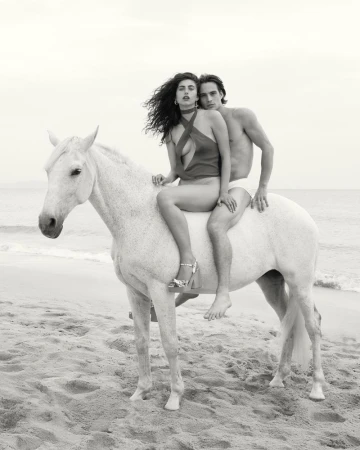 Black and white portrait of an adult couple riding a white horse on a sandy beach with the ocean and sky in the background.