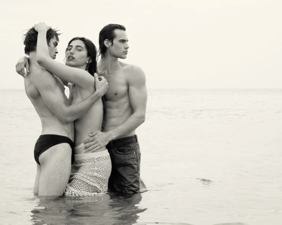 Three shirtless men, standing close in the water. One in dark swim briefs, one in denim jeans, one in knit crochet skirt. Monochrome.