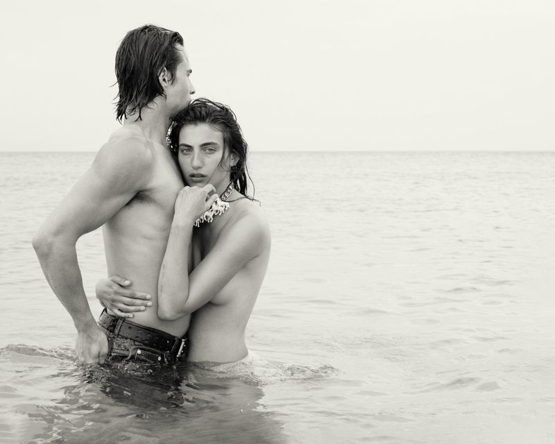 Monochrome portrait of a topless couple standing in ocean water, embracing, with a sandy beach and horizon visible in the background.