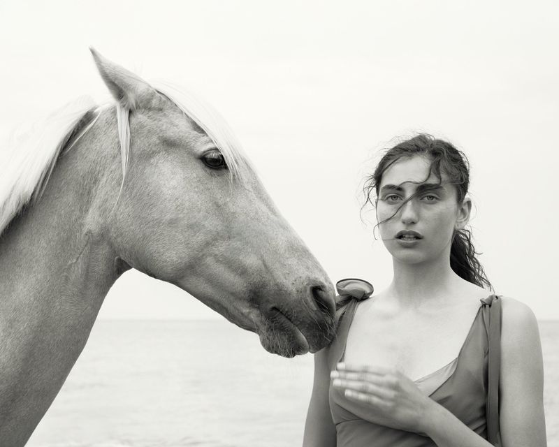 Black and white portrait of a woman in red dress next to a horse head, both near a body of water and a light sky. 