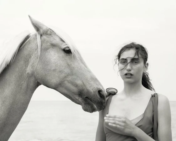 Black and white portrait of a woman in red dress next to a horse head, both near a body of water and a light sky. 