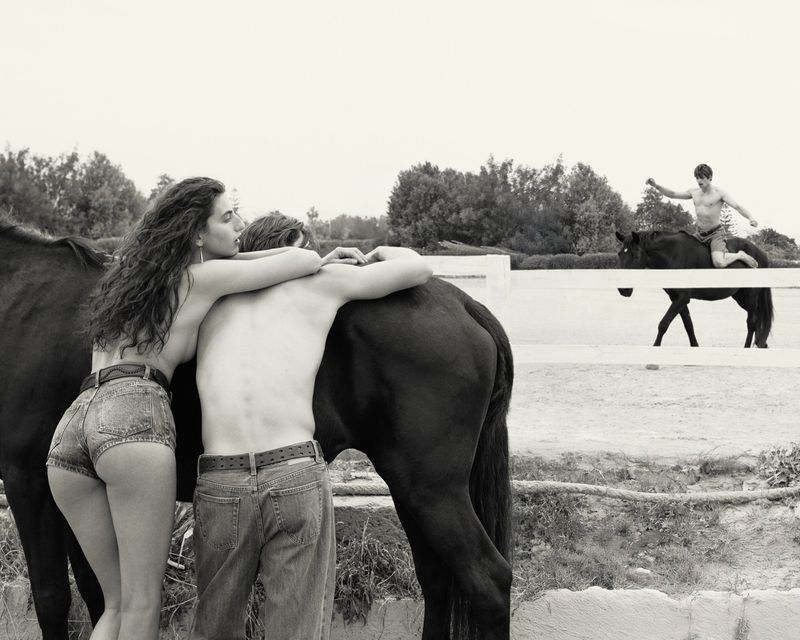 A woman and man embracing near a black horse in an outdoor paddock while another man rides a horse in the background