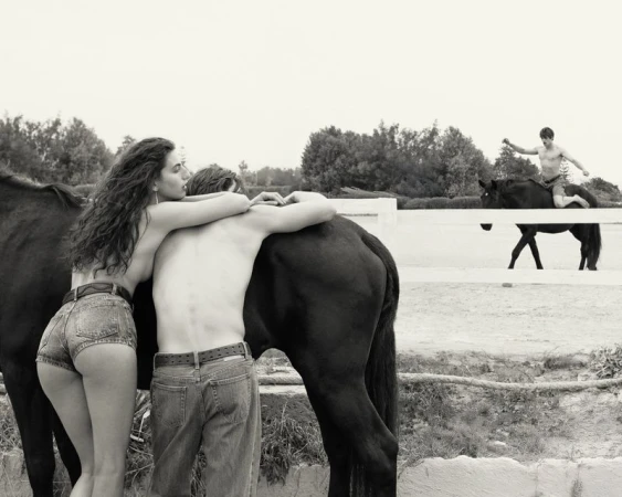 A woman and man embracing near a black horse in an outdoor paddock while another man rides a horse in the background