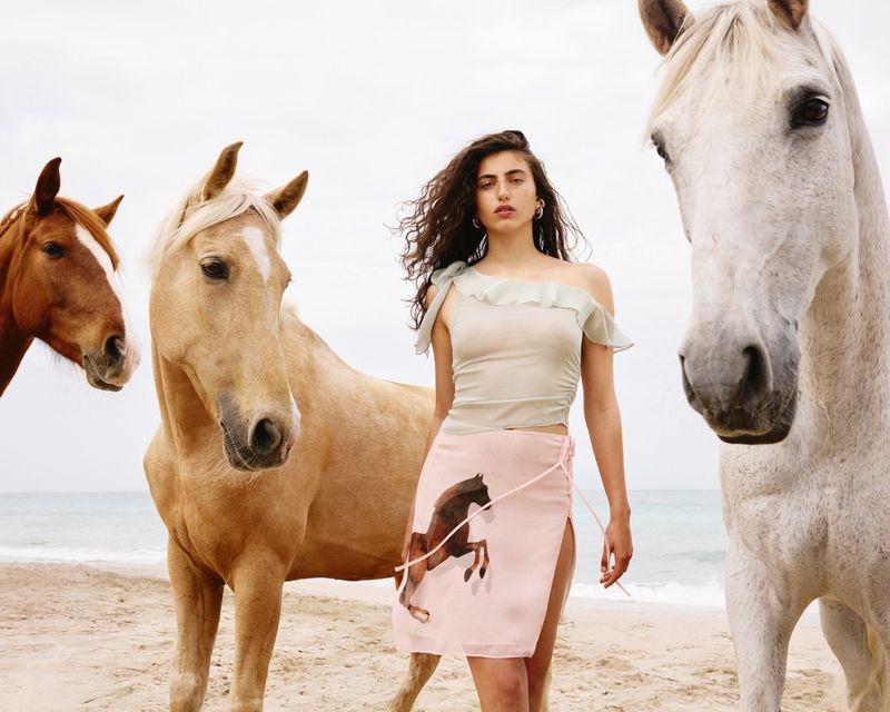Young woman poses with horses on a sandy beach under an overcast sky, modeling a pale outfit with a horse-print skirt.