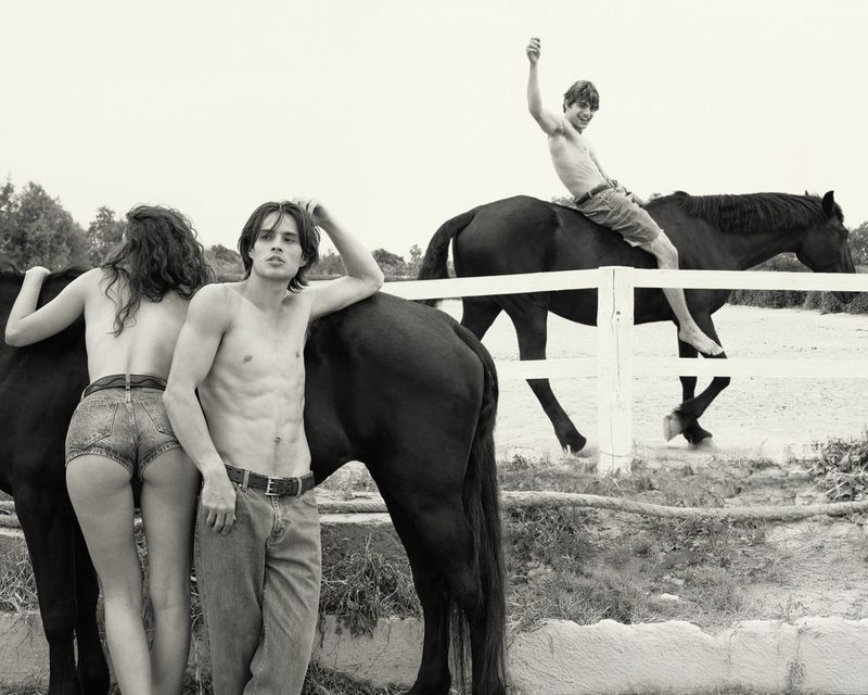 Black and white photo of three shirtless young adults in denim with horses in an outdoor setting with a white fence and trees.