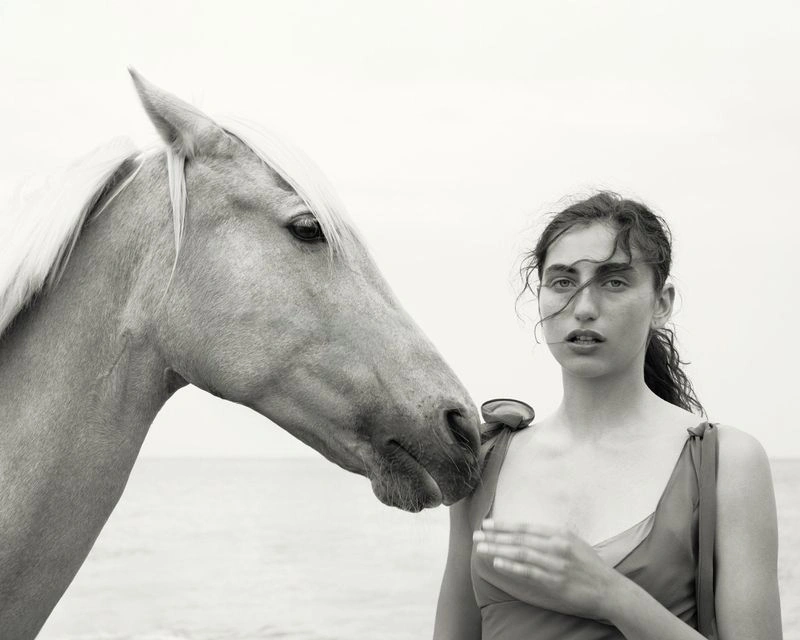 Black and white editorial portrait of a woman standing next to a horse against an open sky background.