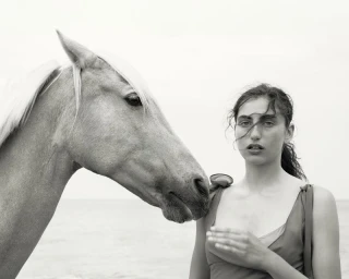 Black and white editorial portrait of a woman standing next to a horse against an open sky background.