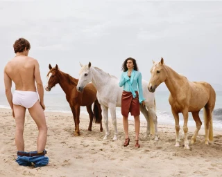 A man in white briefs standing on a sandy beach with horses while a woman stands nearby