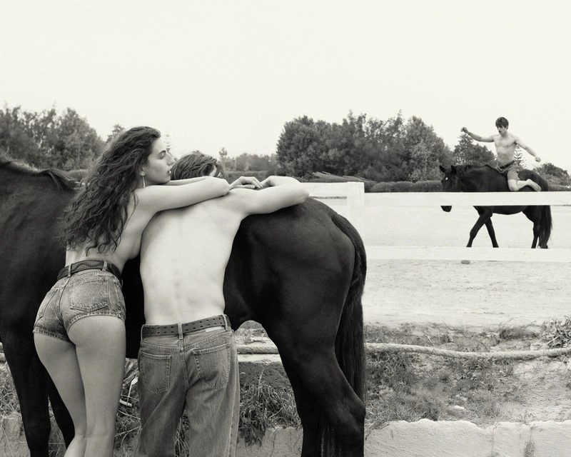 A woman and man embracing near a black horse in an outdoor paddock while another man rides a horse in the background