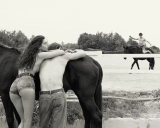 A woman and man embracing near a black horse in an outdoor paddock while another man rides a horse in the background