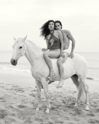 Black and white editorial fashion campaign image by Venroy featuring two models on a white horse on a sandy beach.
