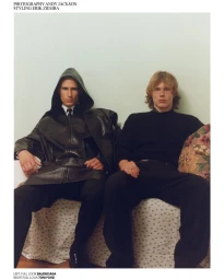 Two male models seated side by side in dark designer menswear against a pale wall for Replica Man Magazine.