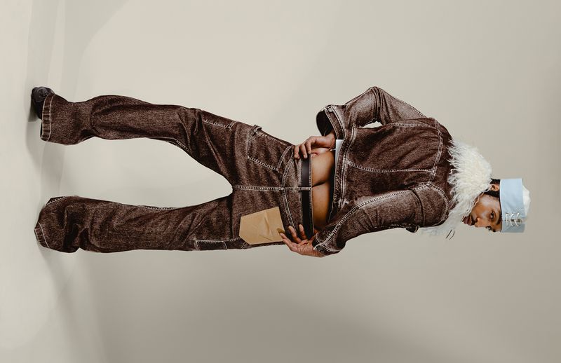 Model Laro Junior posing from behind in a brown denim jacket with fur trim and matching flared trousers against a studio backdrop.