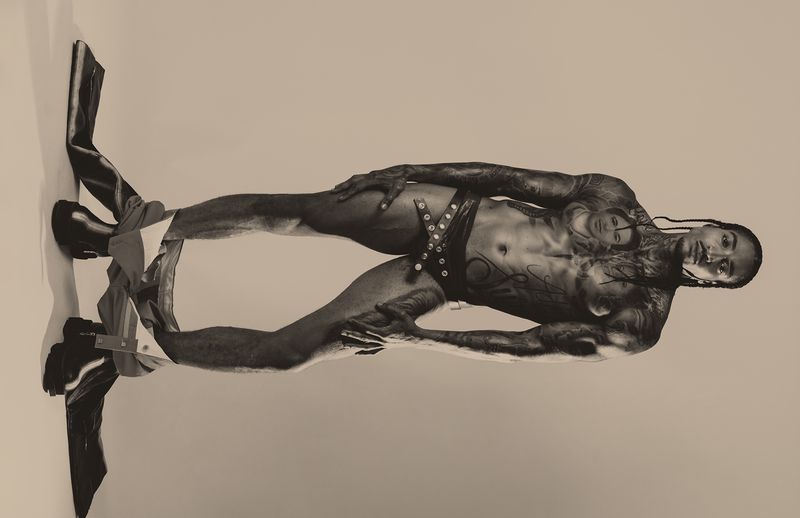Model Laro Junior poses in studded black briefs and boots with trousers lowered against a warm neutral studio backdrop.