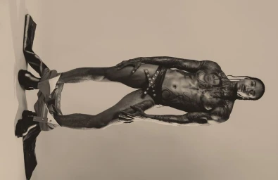 Model Laro Junior poses in studded black briefs and boots with trousers lowered against a warm neutral studio backdrop.