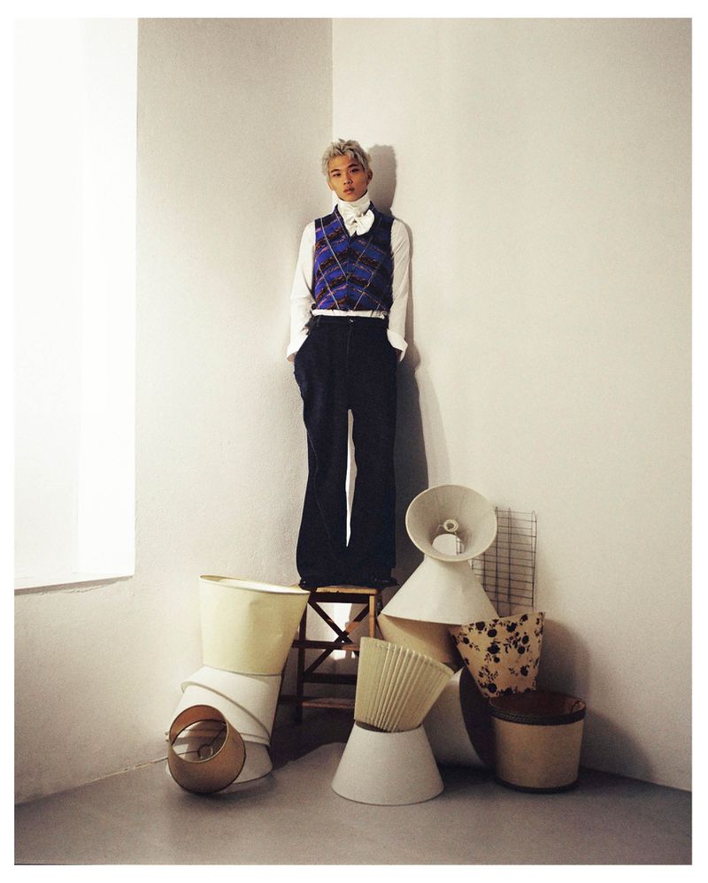 Junseo Kim standing on a wooden stool in a room corner surrounded by stacked lampshades, wearing a blue knit vest and dark trousers.