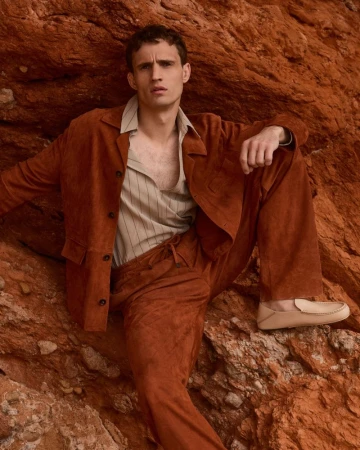 Model Julian Schneyder posing against a red rock wall wearing a rust suede jacket, matching trousers, and cream loafers.