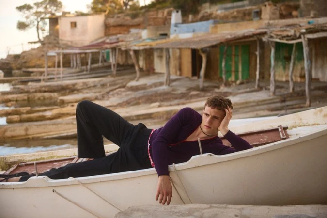 Model Julian Schneyder reclines in a white rowboat wearing a purple knit top and black trousers beside rustic coastal boathouses.