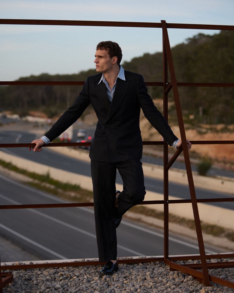 Julian Schneyder leaning against a metal frame structure outdoors, wearing a charcoal double-breasted suit and a striped blue button-down shirt.