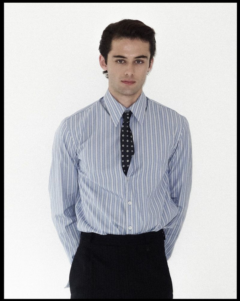Actor and model William Franklyn-Miller posing in a blue striped dress shirt and black tie against a white studio background.