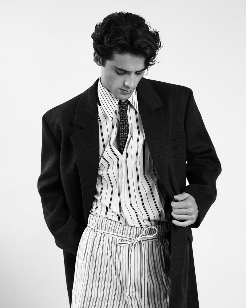 William Franklyn-Miller wearing an oversized dark coat and striped trousers against a white seamless background.