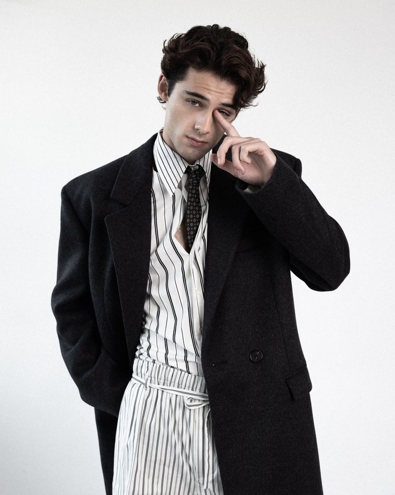 William Franklyn-Miller poses in an oversized black coat, striped collared shirt, and tie against a white studio backdrop.
