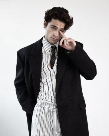 William Franklyn-Miller poses in an oversized black coat, striped collared shirt, and tie against a white studio backdrop.