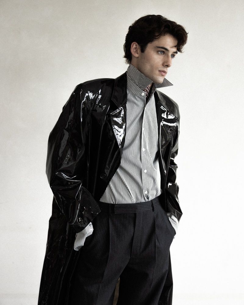 William Franklyn-Miller wearing a glossy black trench coat and striped collared shirt against a white backdrop