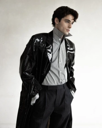 William Franklyn-Miller wearing a glossy black trench coat and striped collared shirt against a white backdrop