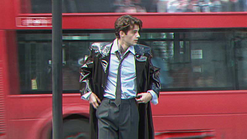 William Franklyn-Miller wearing a glossy black coat, dress shirt, and tie standing in front of a red double-decker bus.