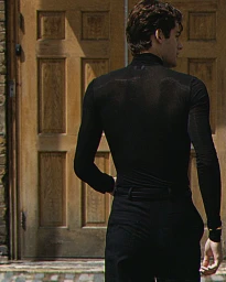 William Franklyn-Miller seen from behind wearing a fitted black long-sleeve top and trousers in front of a wooden door.