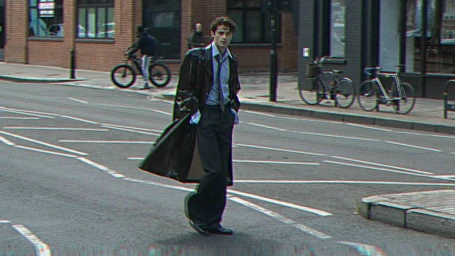 William Franklyn-Miller walking across an urban street wearing an open dark trench coat, blue shirt, and tie with CRT scanline effect.