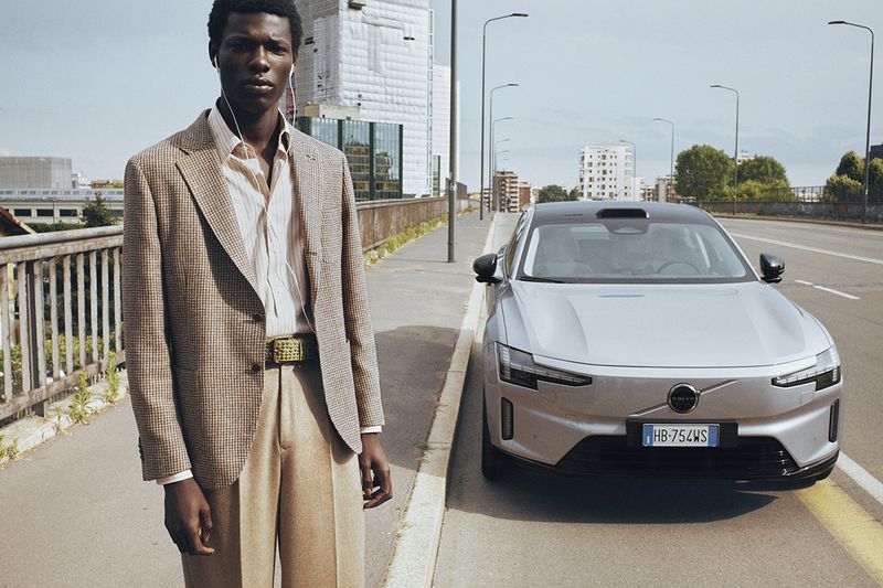 A Black man in a houndstooth blazer and tan trousers stands next to a silver Volvo SUV on an urban bridge.