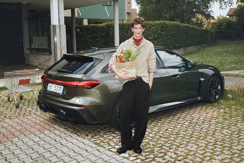 Man holding a brown paper grocery bag with vegetables while standing next to a dark green station wagon in a parking area.