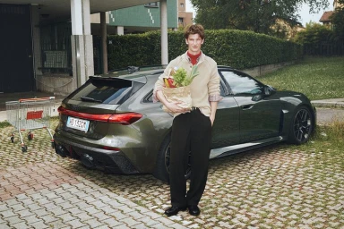 Man holding a brown paper grocery bag with vegetables while standing next to a dark green station wagon in a parking area.