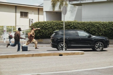 Two men running across a paved street with rolling luggage toward a black SUV parked next to a green hedge.