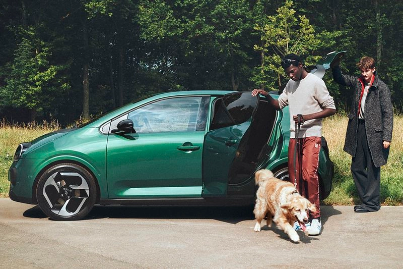 Two men standing next to a green car outdoors with a golden retriever dog on a paved road near a grassy field and trees