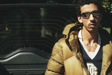 Man wearing sunglasses and an open tan puffer jacket over a navy sweater, posed against a dark vehicle tailgate.