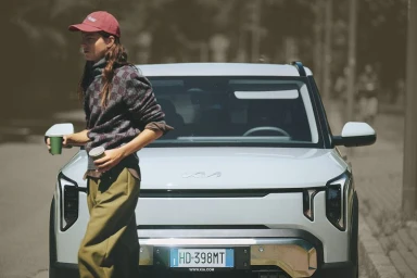 Model in a red cap and patterned knit sweater holding takeaway coffee cups while walking in front of a white SUV on a street.