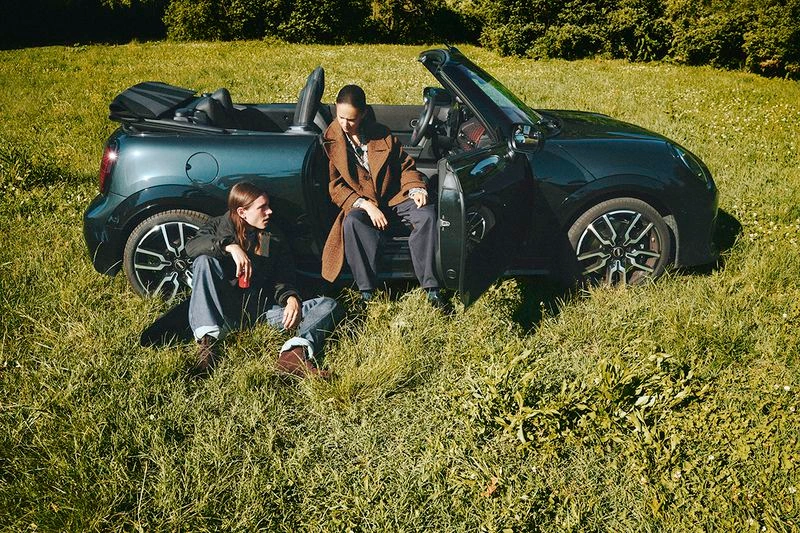 Two fashion models posing by a dark green convertible car parked in a sunlit grassy field.