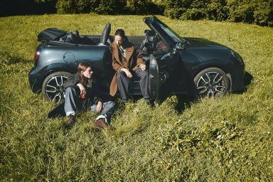 Two fashion models posing by a dark green convertible car parked in a sunlit grassy field.