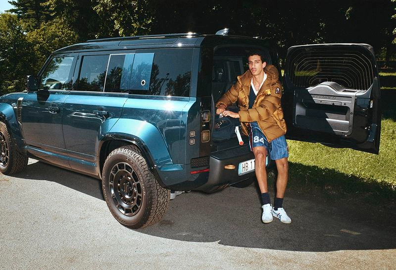 Man in a brown puffer jacket and athletic shorts seated in the open rear trunk of a teal SUV parked beside grassy trees.