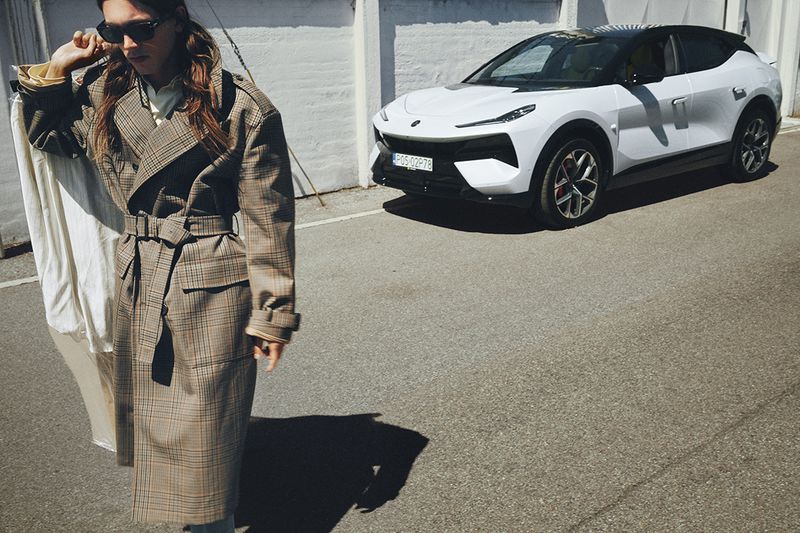 Man in dark sunglasses wearing a belted plaid trench coat walking along an asphalt road with a white SUV in the background.
